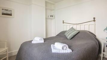 Ein ordentlich bezogenes Bett mit einer grauen Steudecke und zwei aufgerollten Handtüchern, ein weißer Nachttisch mit einer Lampe und ein weißer Kleiderschrank im Hintergrund.