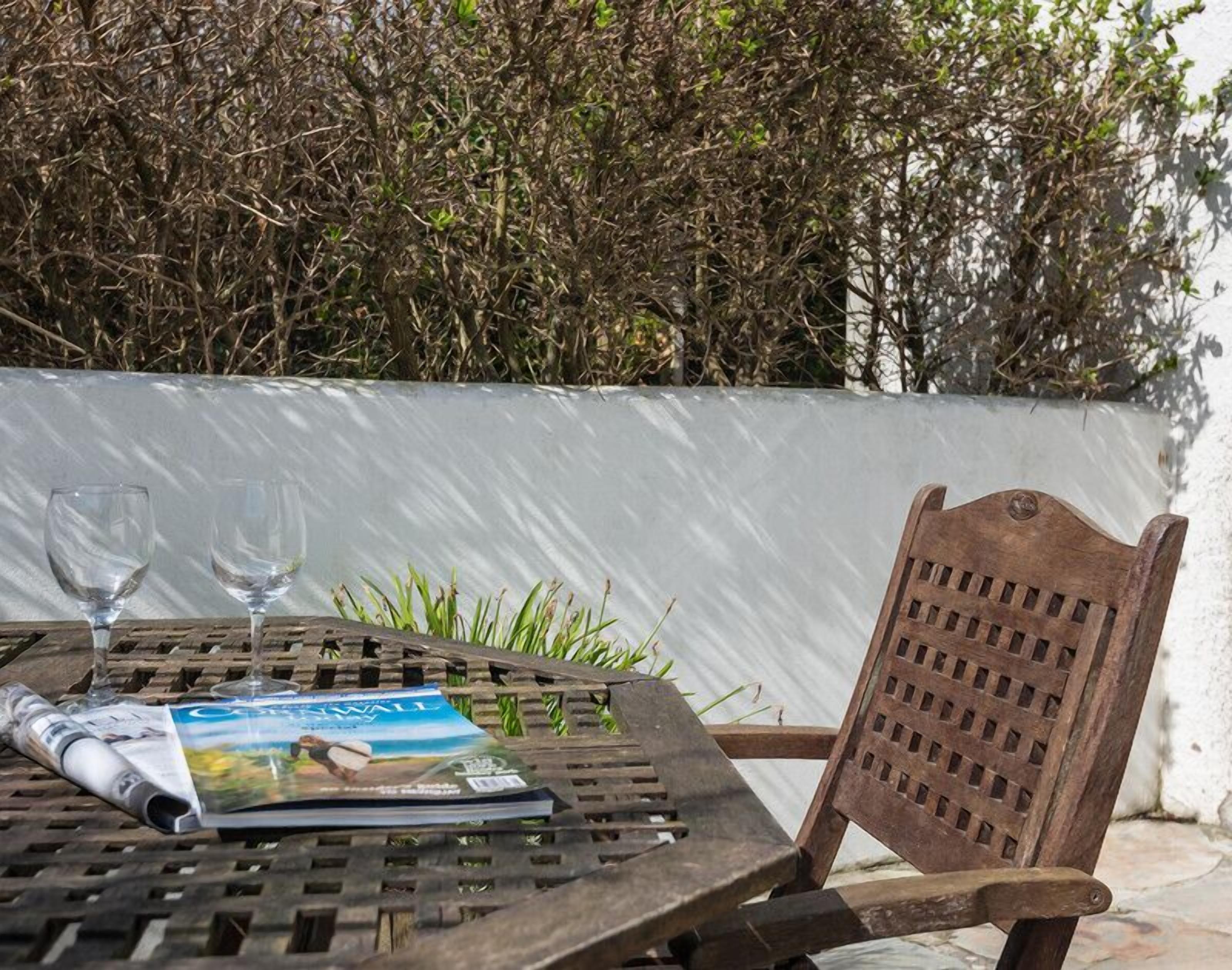 Une table en bois avec deux verres et un magazine, une chaise en bois, et un mur blanc avec de la verdure en arrière-plan.