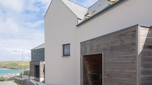 Una casa moderna de dos pisos con un muro de contención de piedra, una puerta de garaje de madera y vista al mar.