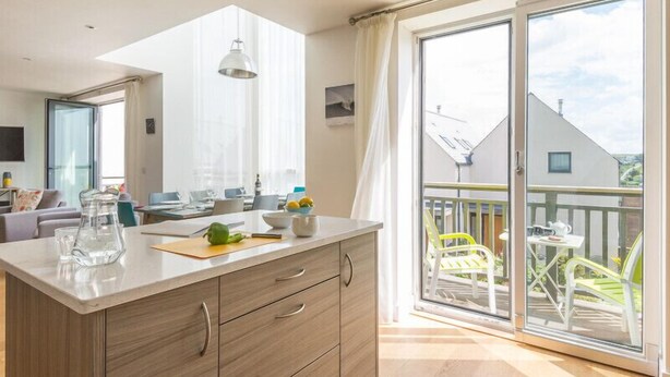 Una cocina moderna con una isla grande, un comedor con mesa y sillas, y un balcón con mobiliario exterior.