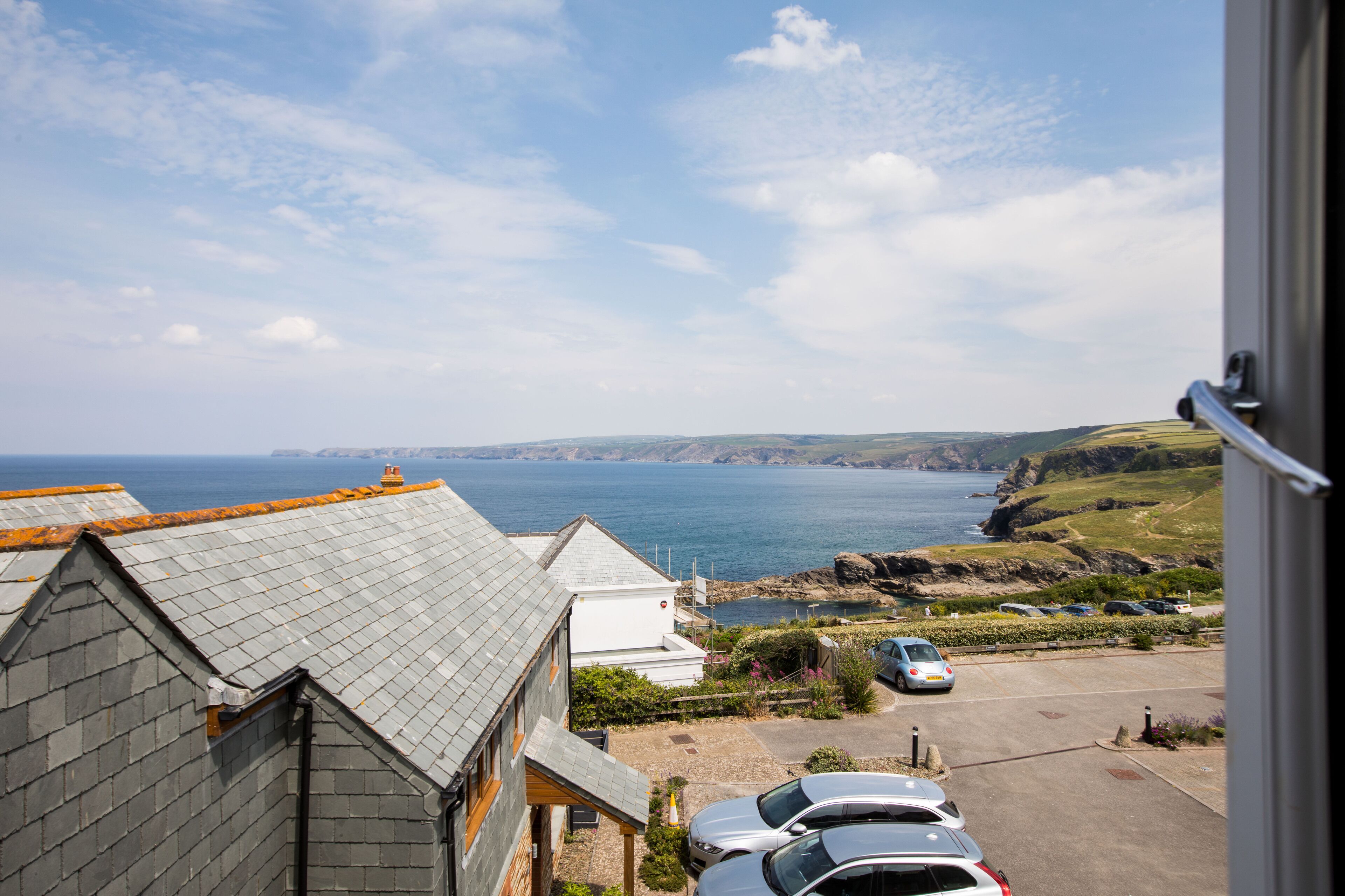A coastal view from a building with a grey roof, a parking area with cars, and a clear sky.