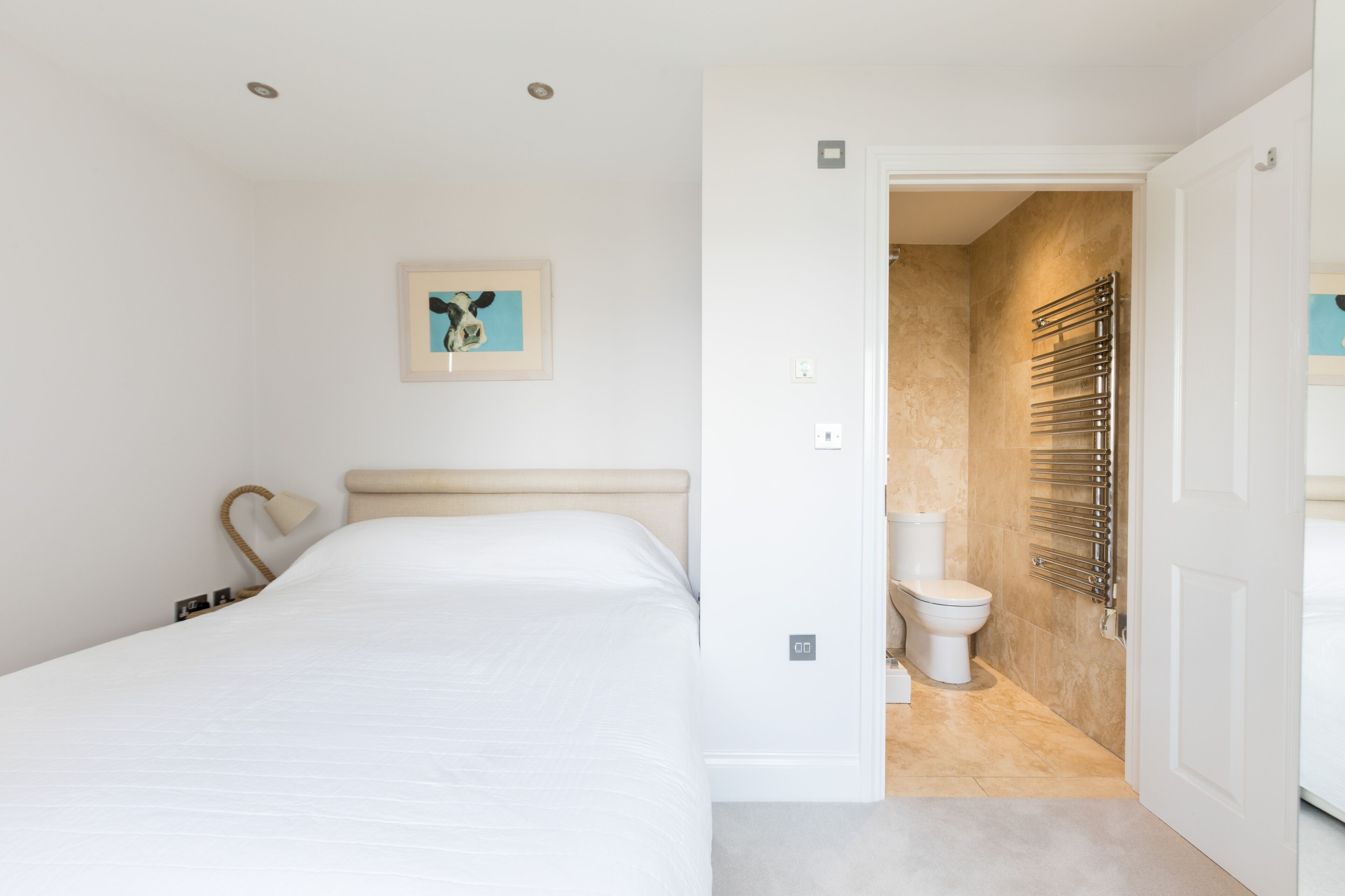 Une chambre à coucher moderne avec un lit, un tableau encadré au mur et une salle de bain visible à travers une porte ouverte.