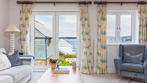 A living room with a sofa, a coffee table, a patterned armchair, and a view through a glass door to a garden. - Rock Lobster (Port Isaac)