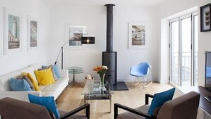 Een moderne woonkamer met een bank, twee fauteuils, een glazen salontafel, een zwarte open haard, en ingelijste foto’s aan de muren.