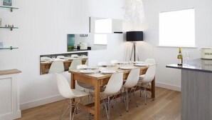 Een moderne eetruimte met een houten tafel en witte stoelen, een kroonluchter en een zichtbare keuken door een spiegel.