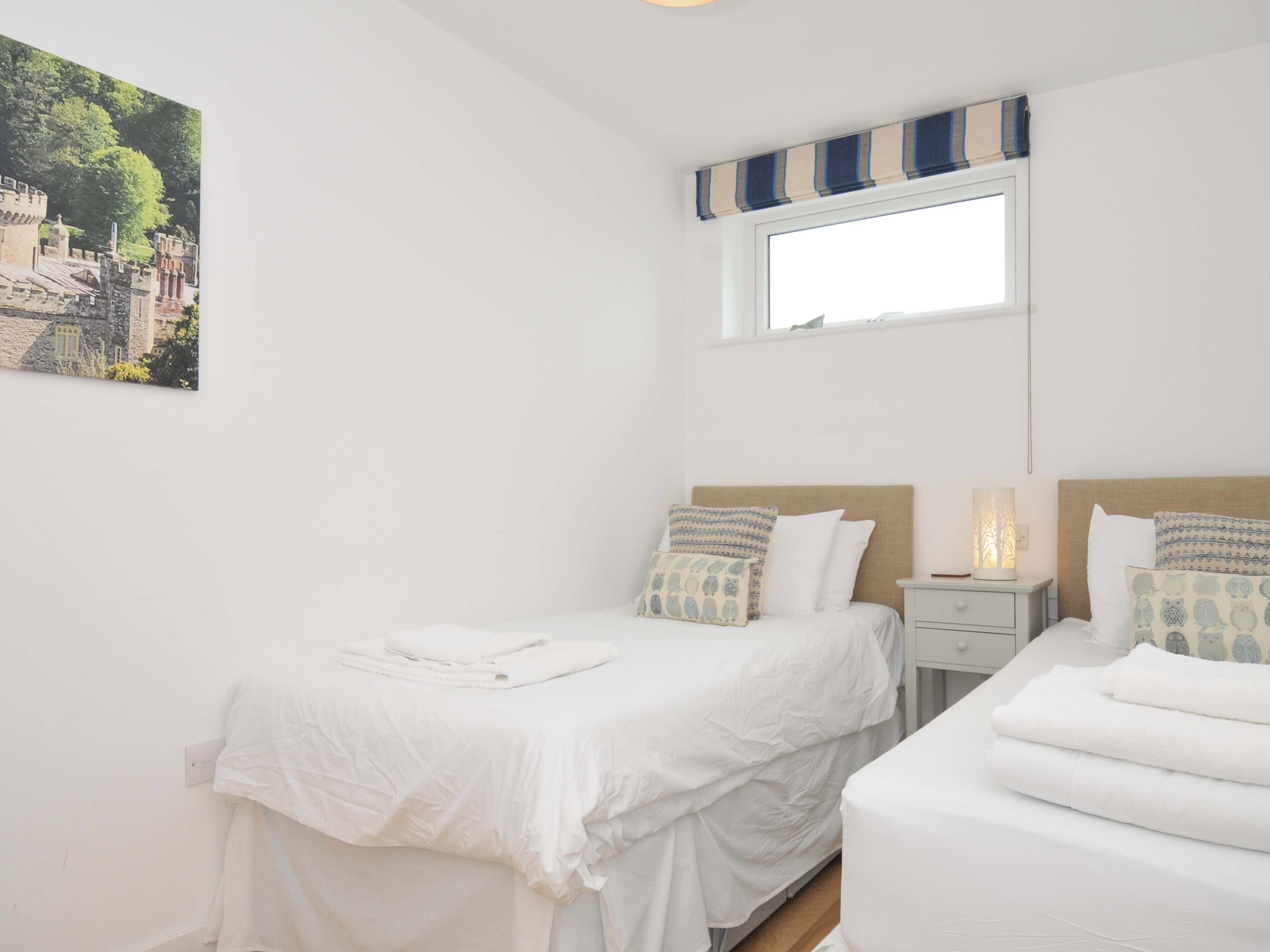 A bedroom with two beds, a window with striped curtains, a nightstand with a lamp, and a painting on the wall.
