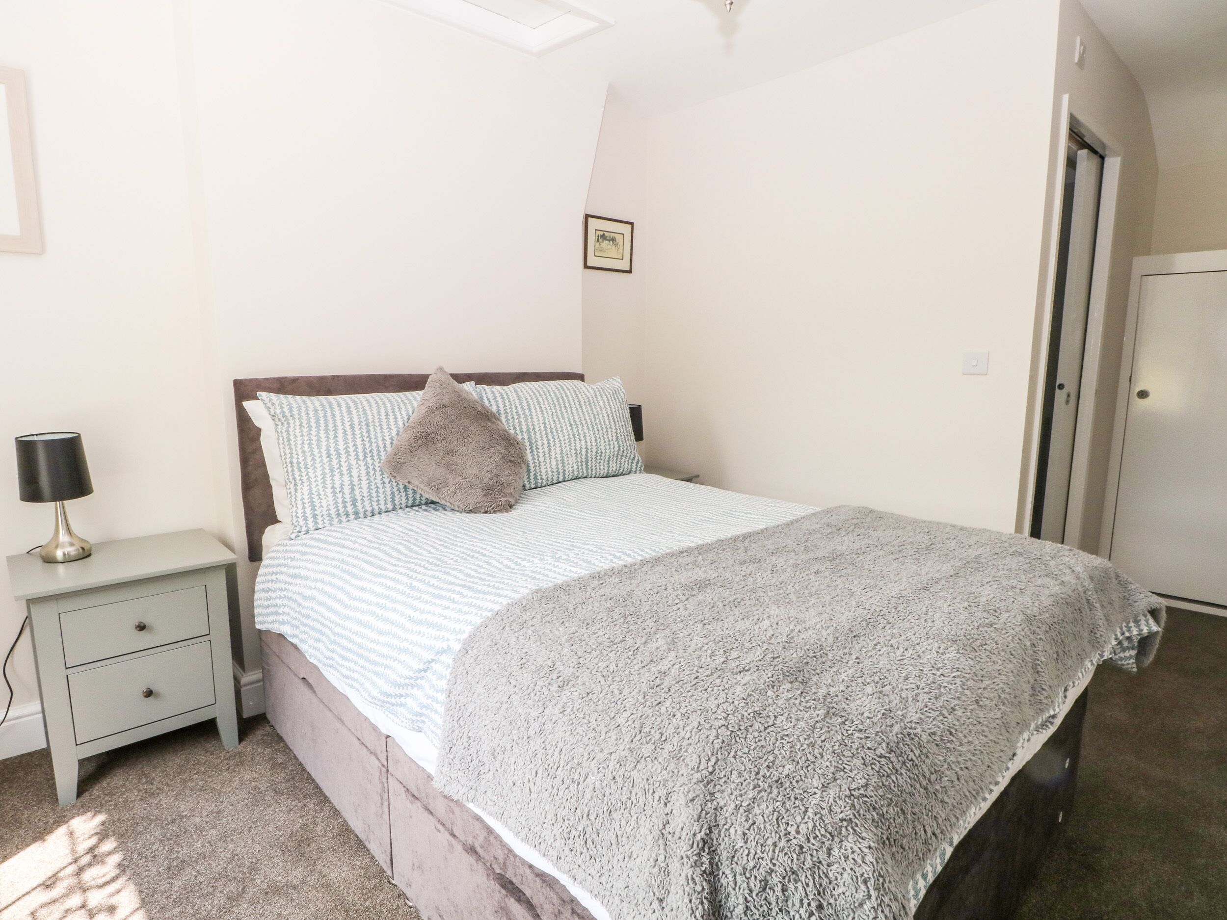Un dormitorio con una cama, una mesita de noche, una lámpara, una puerta y un cuadro pequeño en la pared.