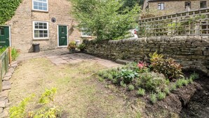 Una casa de piedra con puerta verde, chimenea de ladrillo y jardín con diversas plantas.