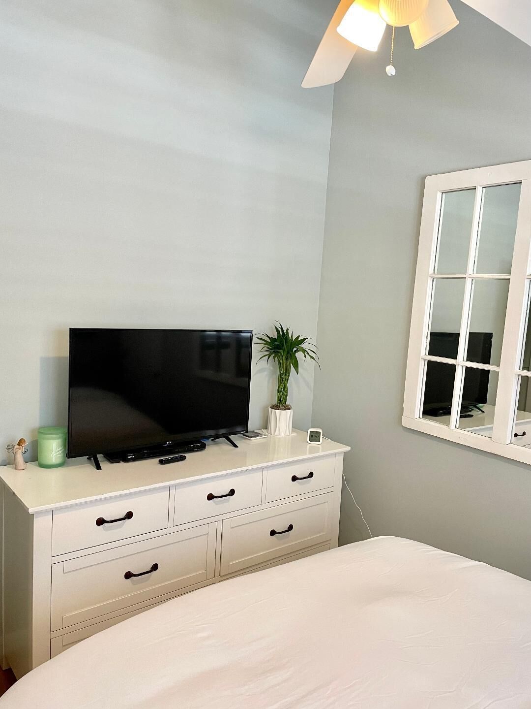 A bedroom with a bed, a white dresser with drawers, a flat-screen TV, a small potted plant, and a window with a white frame.