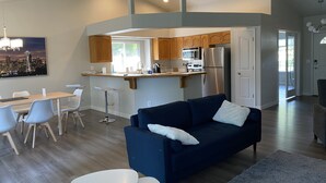 A modern living room with a dark blue sofa, a white coffee table, and a dining area with a white table and chairs. A kitchen with wooden cabinets and stainless steel appliances is visible in the background. - Just like home, clean in great family friendly location (Boise)