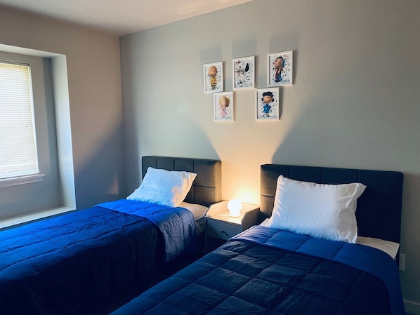 A room with two beds, each with a headboard and a bedside table with a lamp. There are framed pictures on the wall above the beds. - Just like home, clean in great family friendly location (Boise)