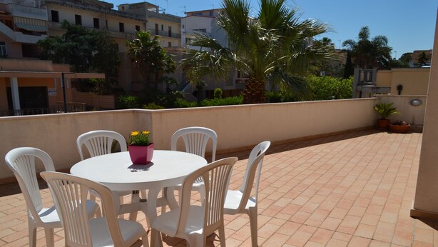 Una mesa redonda con seis sillas blancas y un florero rosa en un patio con baldosas.