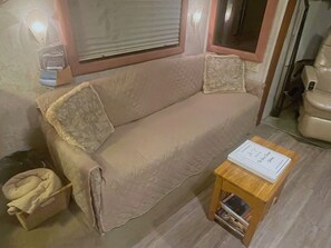Un sofá color beige con funda acolchada, una mesita de madera con un libro, y una ventana con persianas.