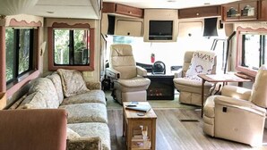 A cozy RV interior with a sofa, two recliners, a small table, and a TV. - The Great Escape - Shirley (Sooke)