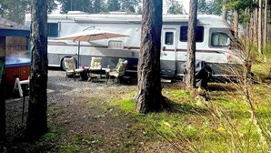 A recreational vehicle (RV) parked in a wooded area with a patio setup including chairs and an umbrella. - The Great Escape - Shirley (Sooke)