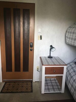 Una habitación con puerta de madera, una mesita de noche con lámpara y una cama con manta a cuadros.