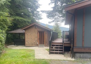 Uma cabana de madeira com varanda e terraço, cercada por árvores e montanhas ao fundo.