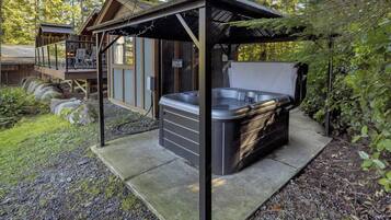 A hot tub under a wooden pergola in a forested area with a wooden deck and a cabin in the background.