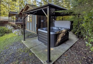 Uma banheira de hidromassagem sob uma pérgola de madeira em área arborizada, com deck de madeira e uma cabana ao fundo.