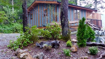 A cabin with a deck, surrounded by trees and a rocky ground.