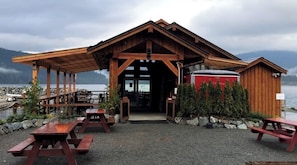 Una baita in legno con un portico, tavoli da picnic e un contenitore rosso, probabilmente per riporre oggetti, vicino a un corso d'acqua.