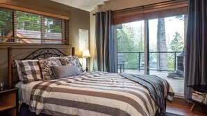 A bedroom with a large bed, a nightstand, a lamp, and a view of the outdoors through a window. - The Great Escape - Port Renfrew (Port Renfrew)