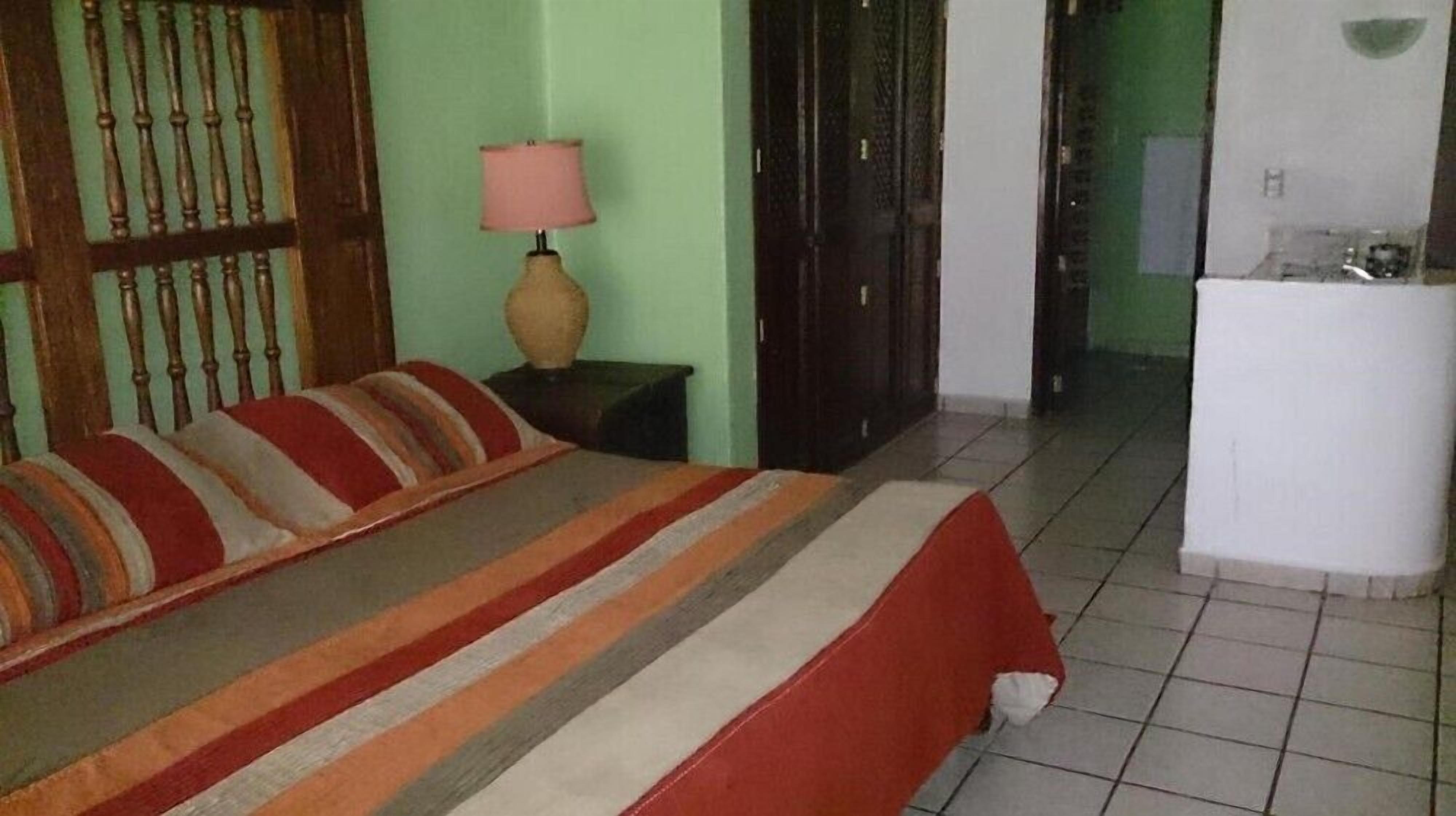 Una cama con cubrecama a rayas, una mesita de noche con lámpara y una puerta que da a otra habitación.