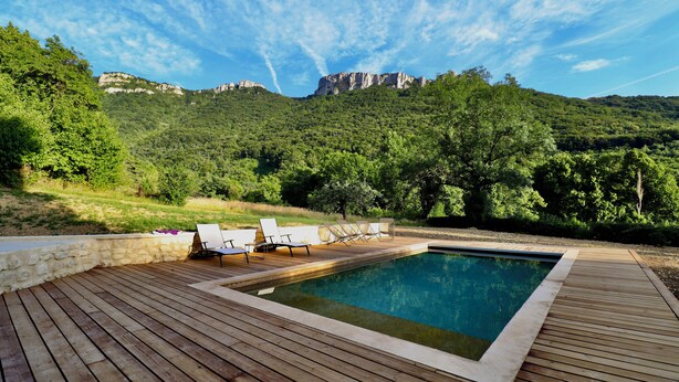 Una piscina rectangular con cubierta de madera y sillas de descanso, rodeada de abundante vegetación y montañas al fondo.