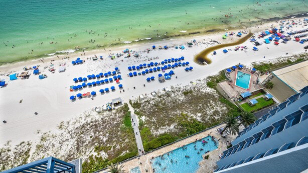 An aerial view of a beach with numerous blue umbrellas, a swimming pool, and a hotel structure. - Beautiful 17th Floor Views! Free Activities + Summer Deals!! (Panama City Beach)