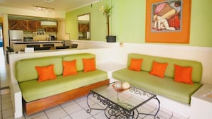 Un espace de vie avec deux canapés verts, une table basse, une cuisine en arrière-plan et un tableau accroché au mur.