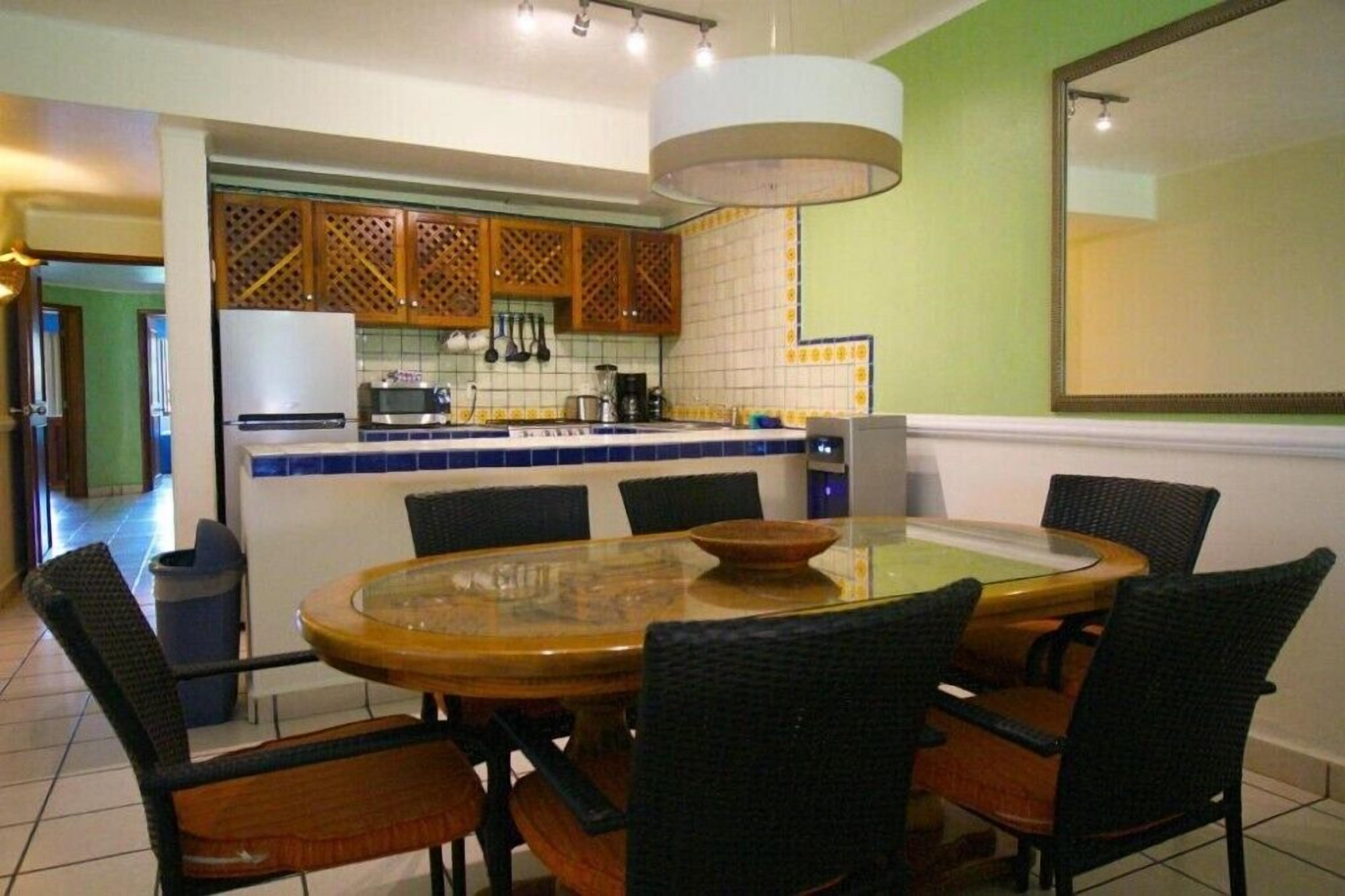 A dining area with a round glass table, black wicker chairs, and a kitchen in the background.