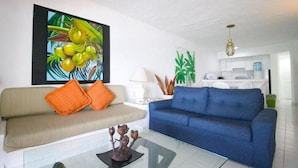 Un soggiorno con un divano beige, un divano blu e un tavolino in vetro. Al muro, un quadro raffigura dei cocco.