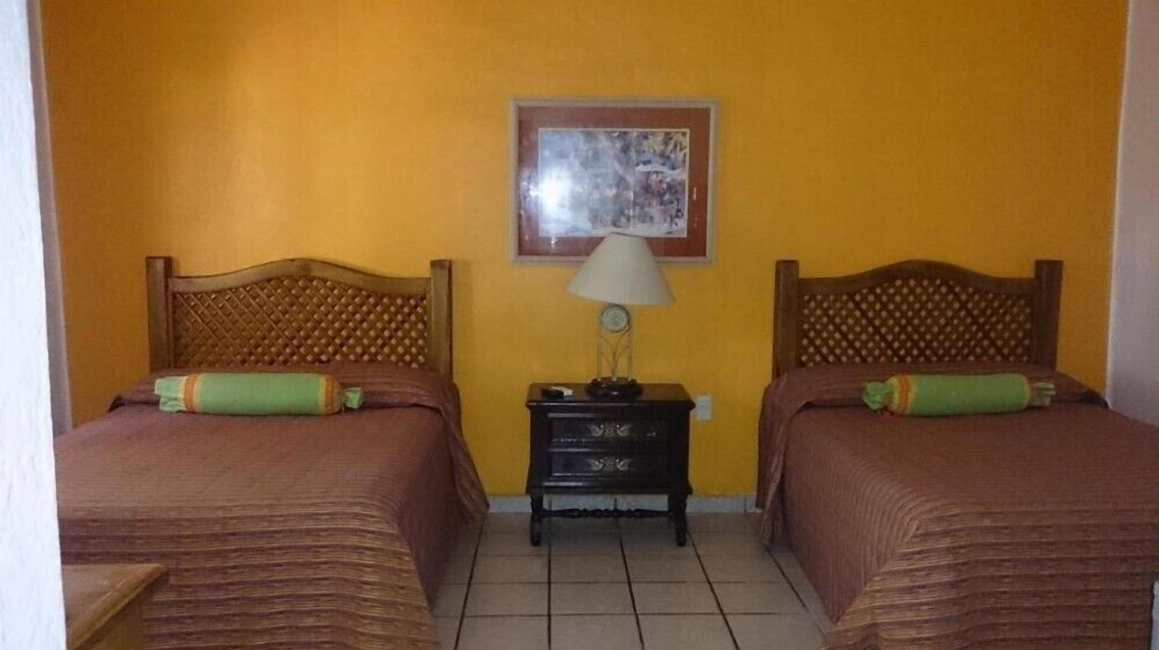 A room with two beds, each with a wooden headboard and a green pillow. There is a nightstand between the beds with a lamp and a clock. The walls are yellow, and there is a framed picture on the wall.