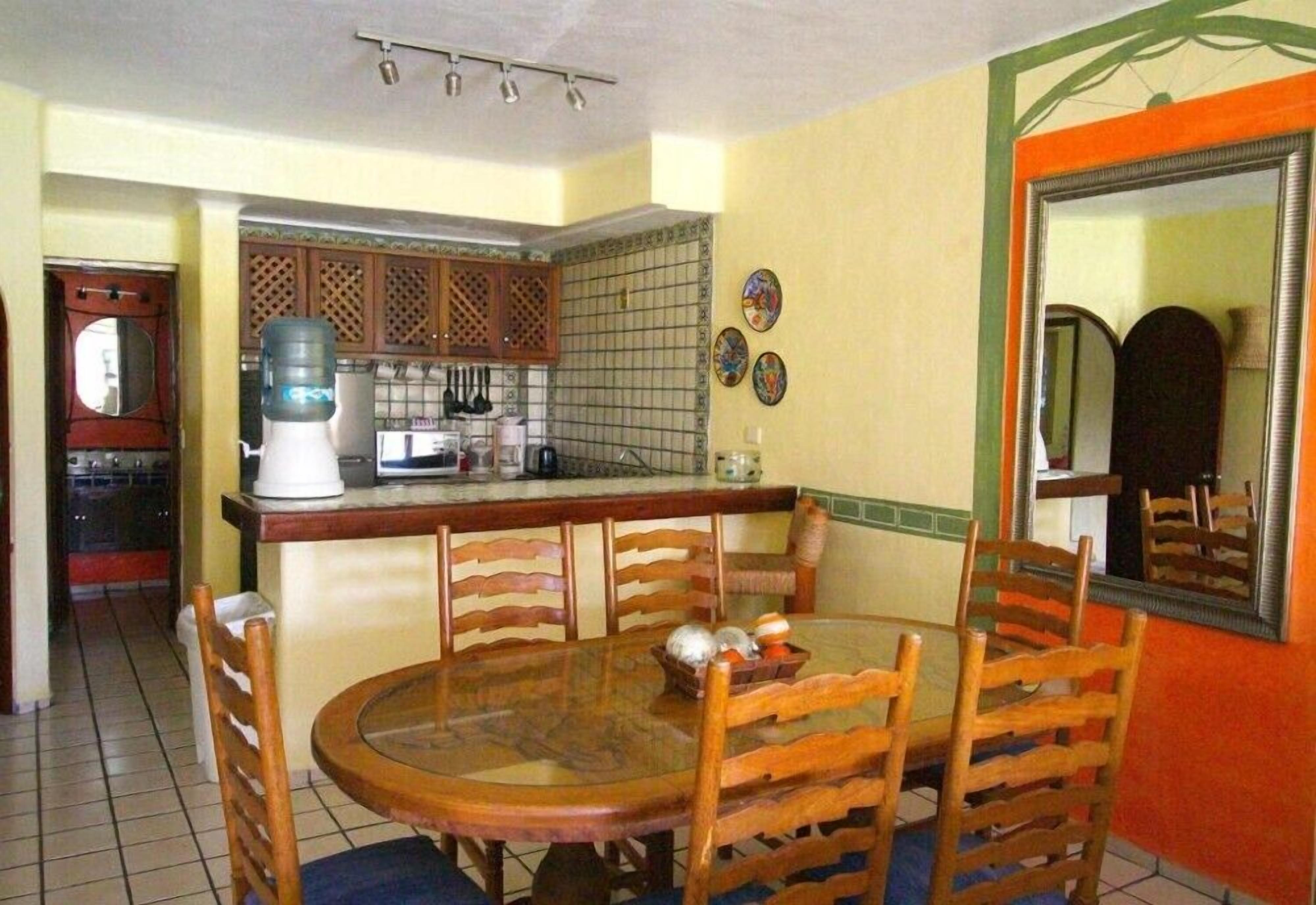 A dining area with a round table and wooden chairs, a kitchen with a tiled backsplash, and a large mirror on the wall.