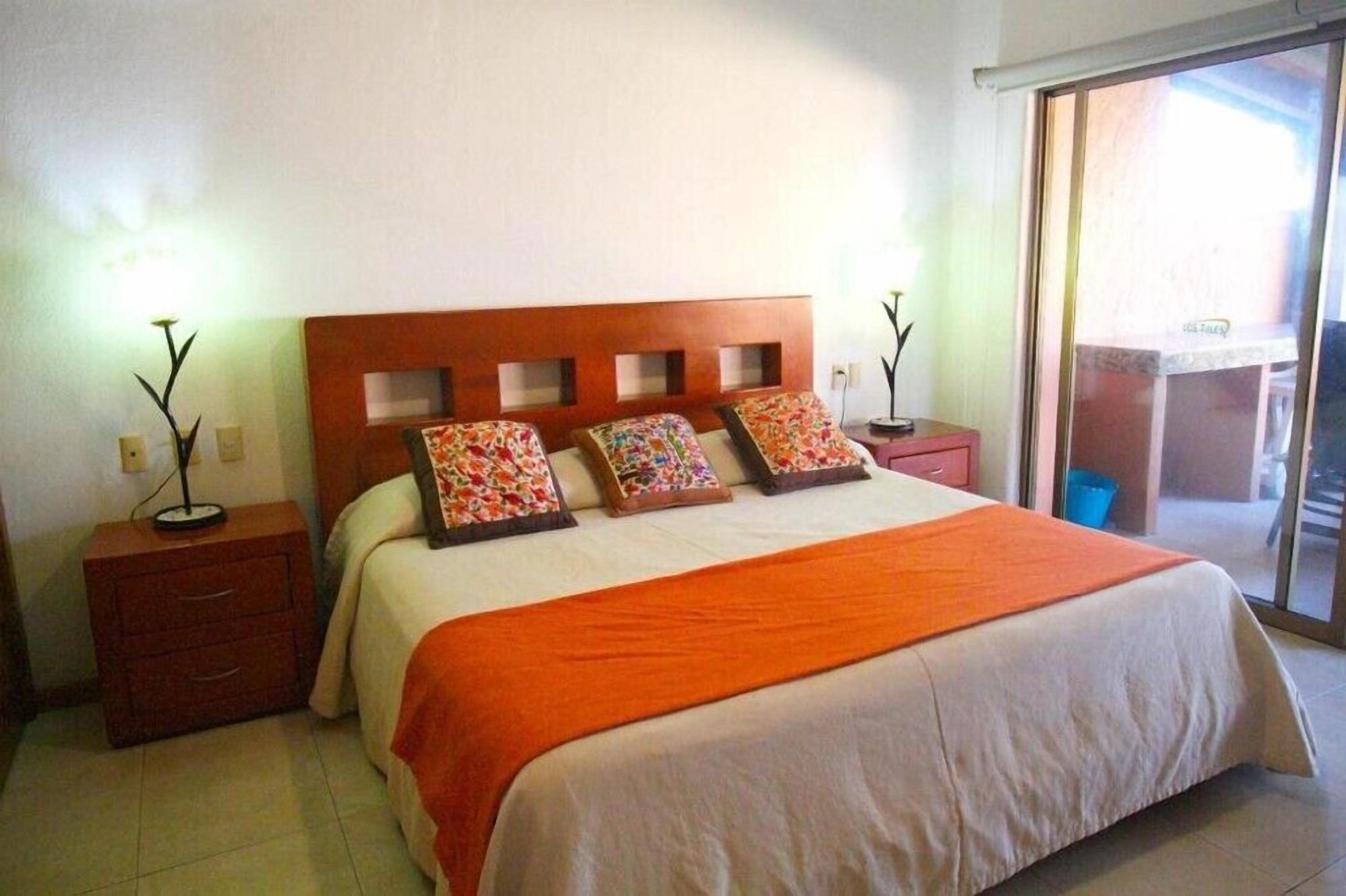 A hotel room with a wooden headboard, a large bed with an orange bedspread, two bedside tables with lamps, and a sliding glass door leading to a balcony.