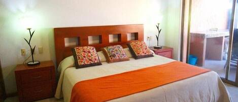 A hotel room with a wooden headboard, a large bed with an orange bedspread, two bedside tables with lamps, and a sliding glass door leading to a balcony.