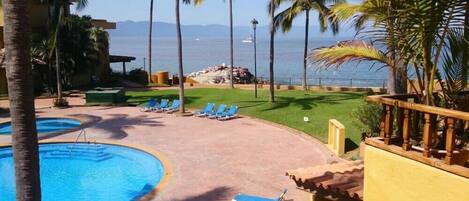 Zona de piscina en un resort con sillas reclinables azules, palmeras y vista al océano.