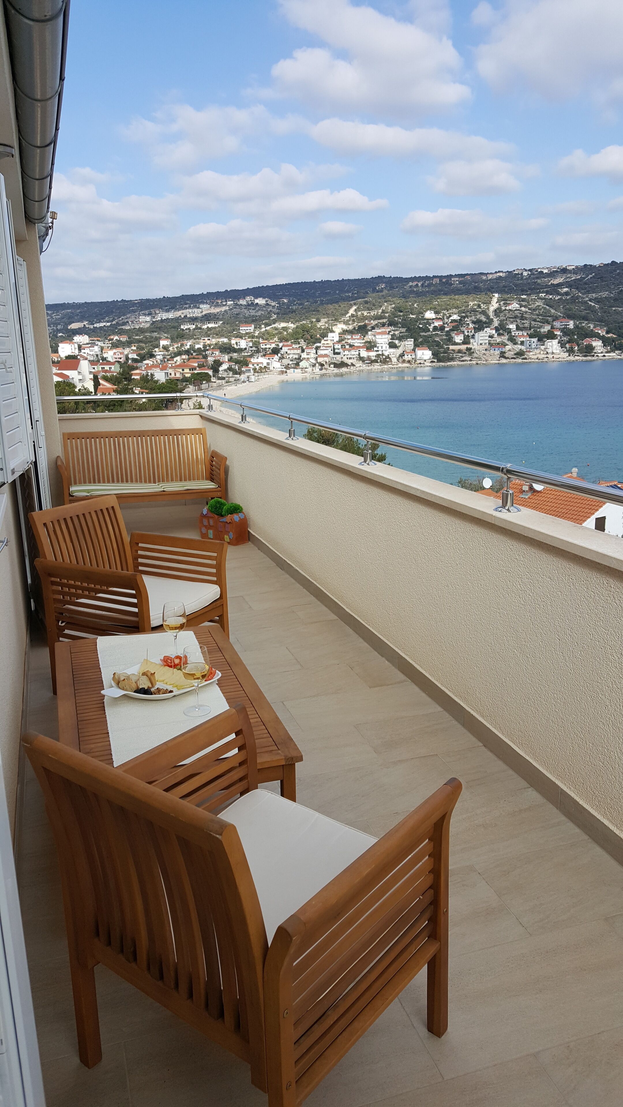 A balcony with wooden furniture and a table set for a meal, overlooking a coastal town and sea.