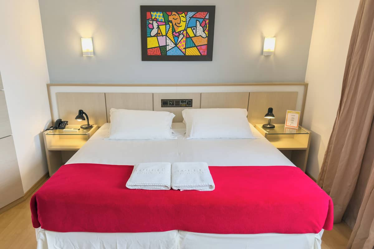 A hotel room with a large bed, two bedside tables, a nightstand, a red blanket, white pillows, and a colorful abstract painting on the wall.