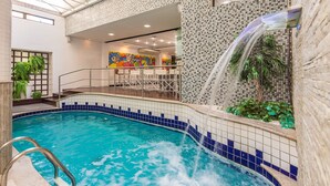 Une piscine intérieure agrémentée d’une cascade, entourée de murs carrelés et d’un espace détente avec des fauteuils.