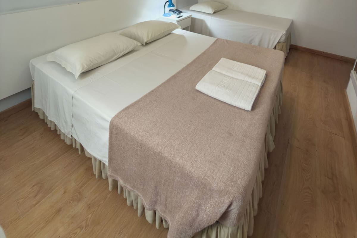 A single-bed room with a wooden floor, a bedside table with a lamp, and a ceiling light fixture.