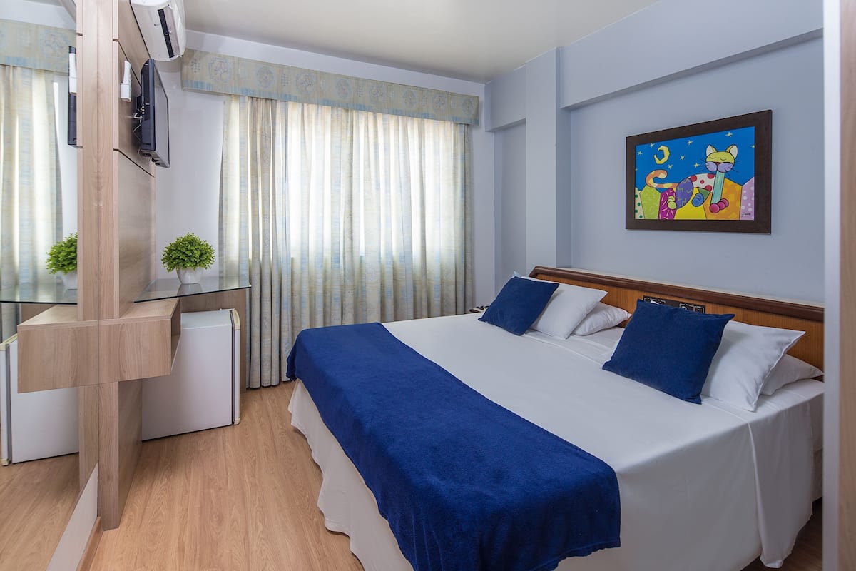 A hotel room with a bed, a desk, a chair, a small table, a potted plant, and a painting on the wall.