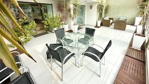 Une terrasse fermée par des parois de verre, avec une table ronde en verre et quatre chaises en osier, entourée de plantes en pot et d’un canapé en osier.