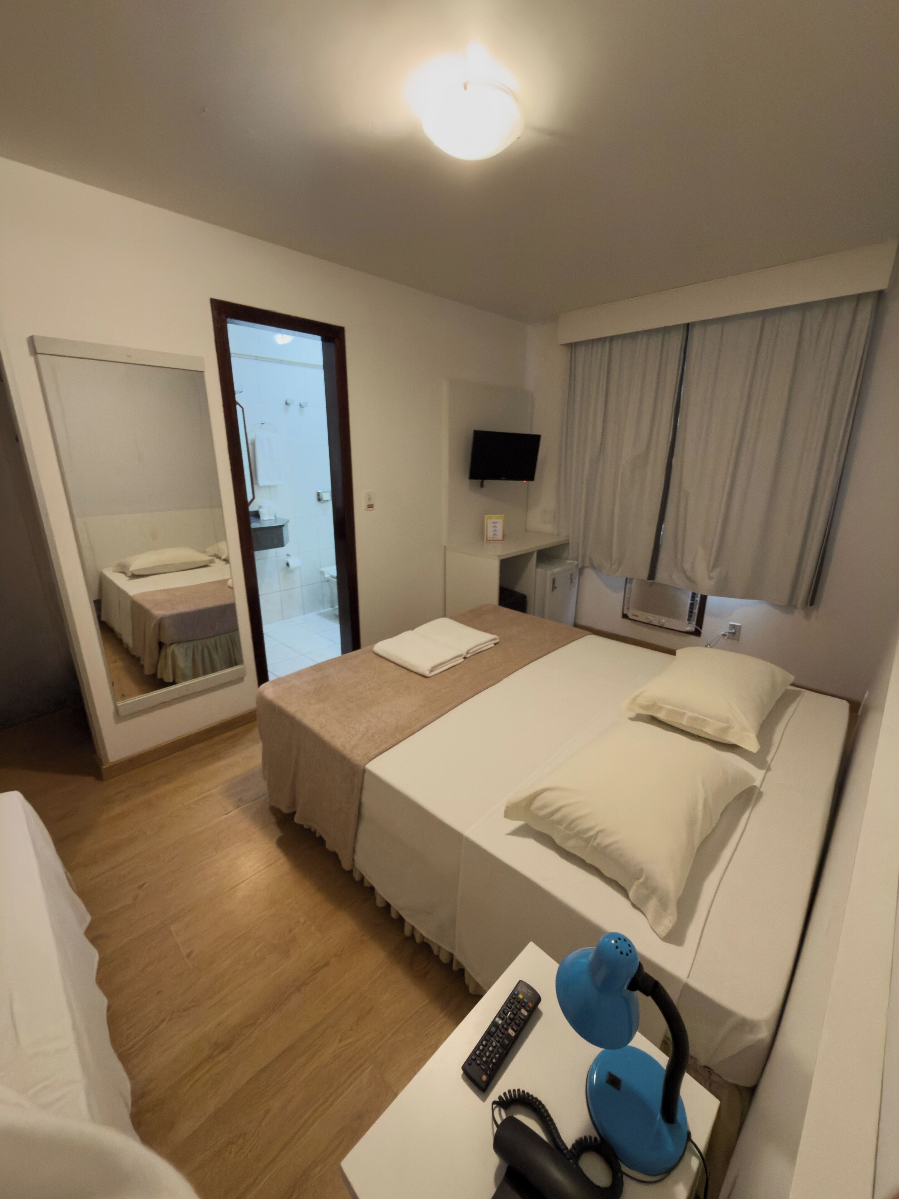A hotel room with a bed, a bedside table with a phone and a lamp, a TV on the wall, and a mirror.