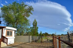 Situated in a fenced area, this mobile home features a front porch with a railing, surrounded by lush vegetation and a clear sky.