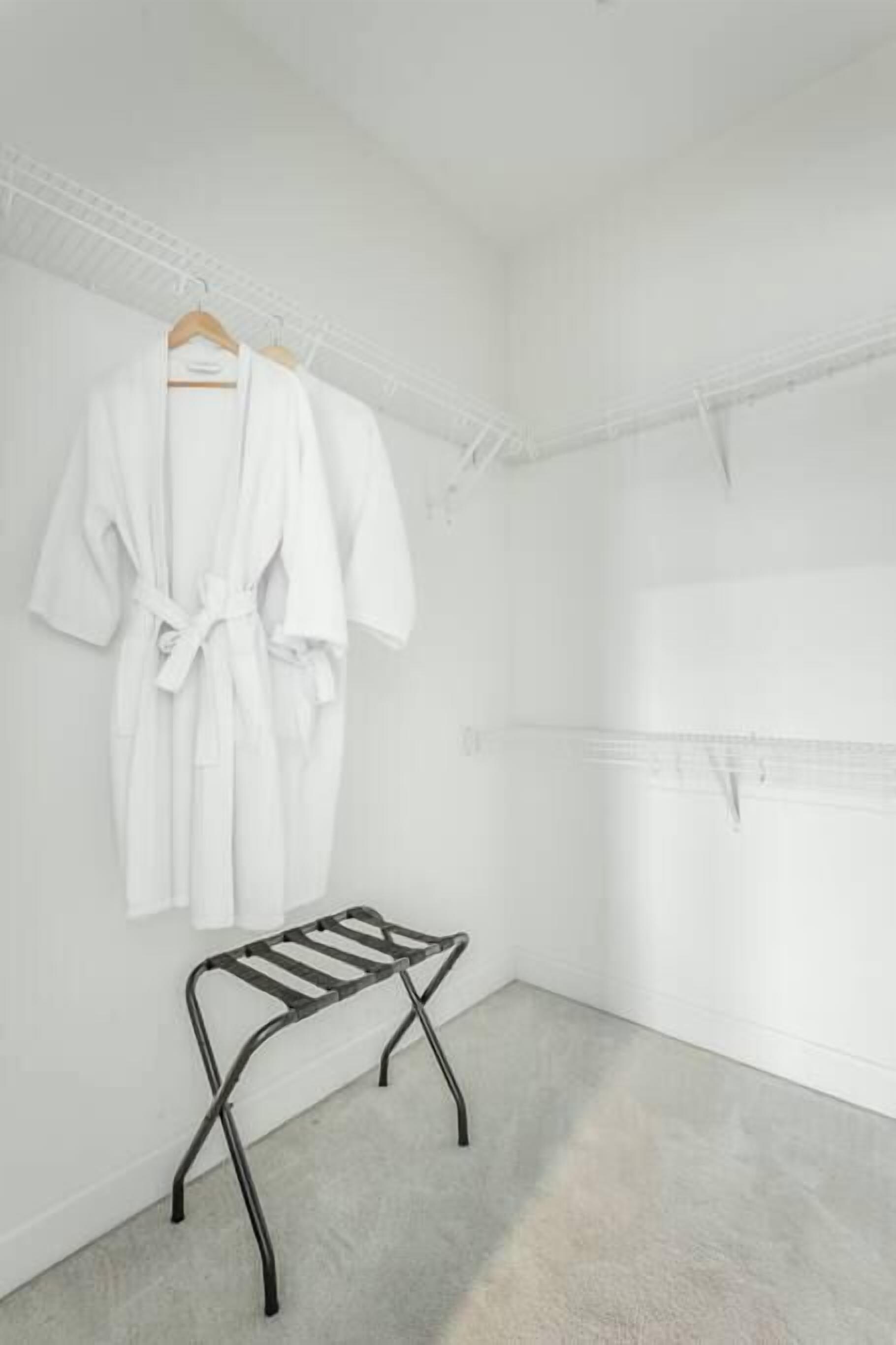 Une pièce blanche contenant un présentoir métallique sur lequel est posée une blouse blanche, un tabouret métallique et une étagère blanche.