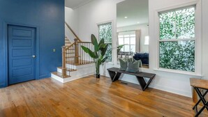 Amplia sala de estar con piso de madera, una puerta azul, una escalera y ventanas grandes con vistas a la vegetación.