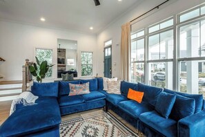 A modern living room with a blue sectional sofa, a large window with a view, and a small kitchen area. - Luxury Reunion House | Walk to Downtown & Stadium (Chattanooga)