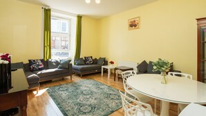 A living room with a sofa, a dining table, and a coffee table. There is a window with curtains, a radiator, and a picture on the wall. - 7 Bedroom Townhouse (Edinburgh)