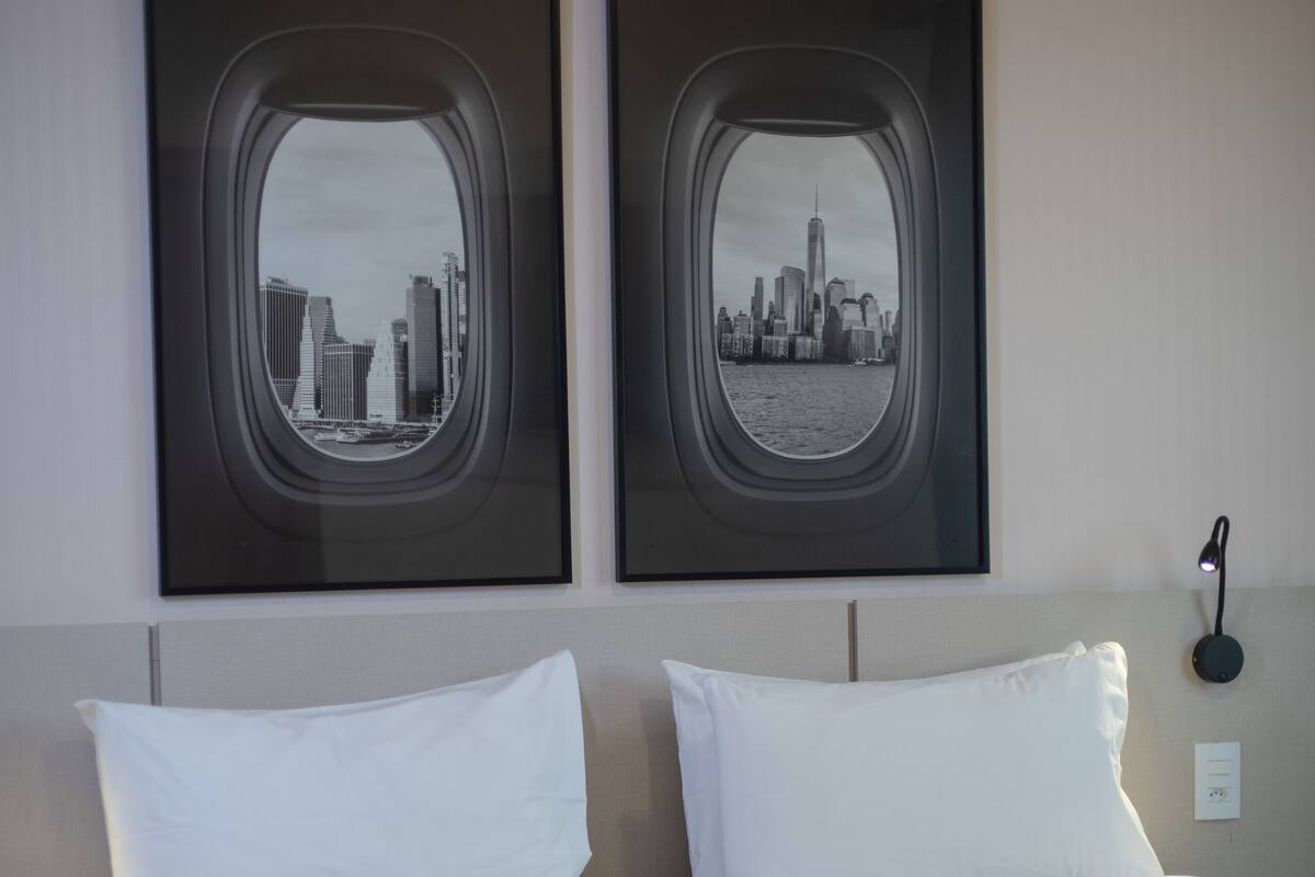 A hotel room with a bed, two framed pictures of cityscapes, a bedside lamp, and an air conditioning unit.